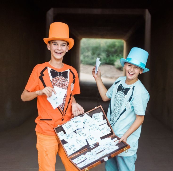 Two teen boys dressed as Harry and Lloyd from Dumb and Dumber in orange and powder blue tuxedo T-shirts and top hats, holding a briefcase full of IOU notes