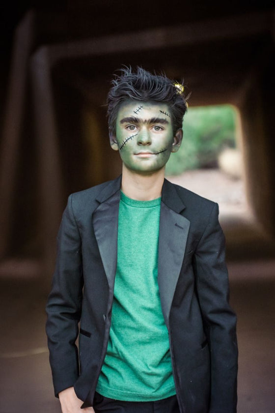 Teen boy dressed as Frankenstein with green face paint, drawn-on stitches, spiked black hair and a black blazer over a green tee