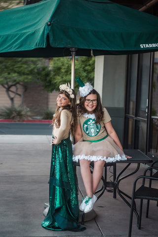 The Frappuccino and Starbucks siren costumes together at a table under a green umbrella