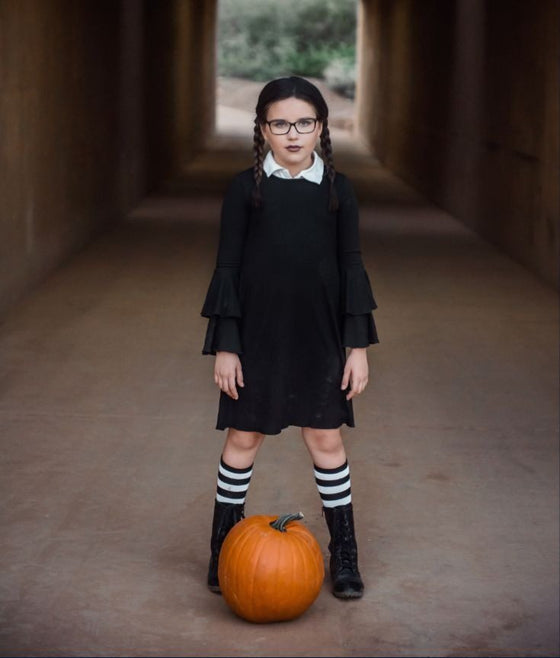 Girl dressed as Wednesday Addams with braids, a black dress with ruffled cuffs and striped socks, standing behind a pumpkin
